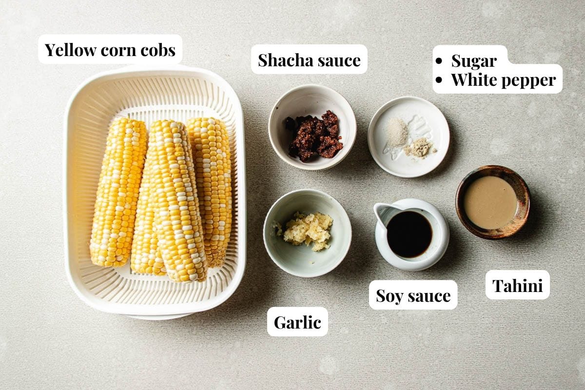 Two yellow corn cobs in a tray, styled Taiwanese street corn–style, with bowls of shacha sauce, sugar, white pepper, tahini, soy sauce, and minced garlic arranged nearby. Ingredients are labeled with text.