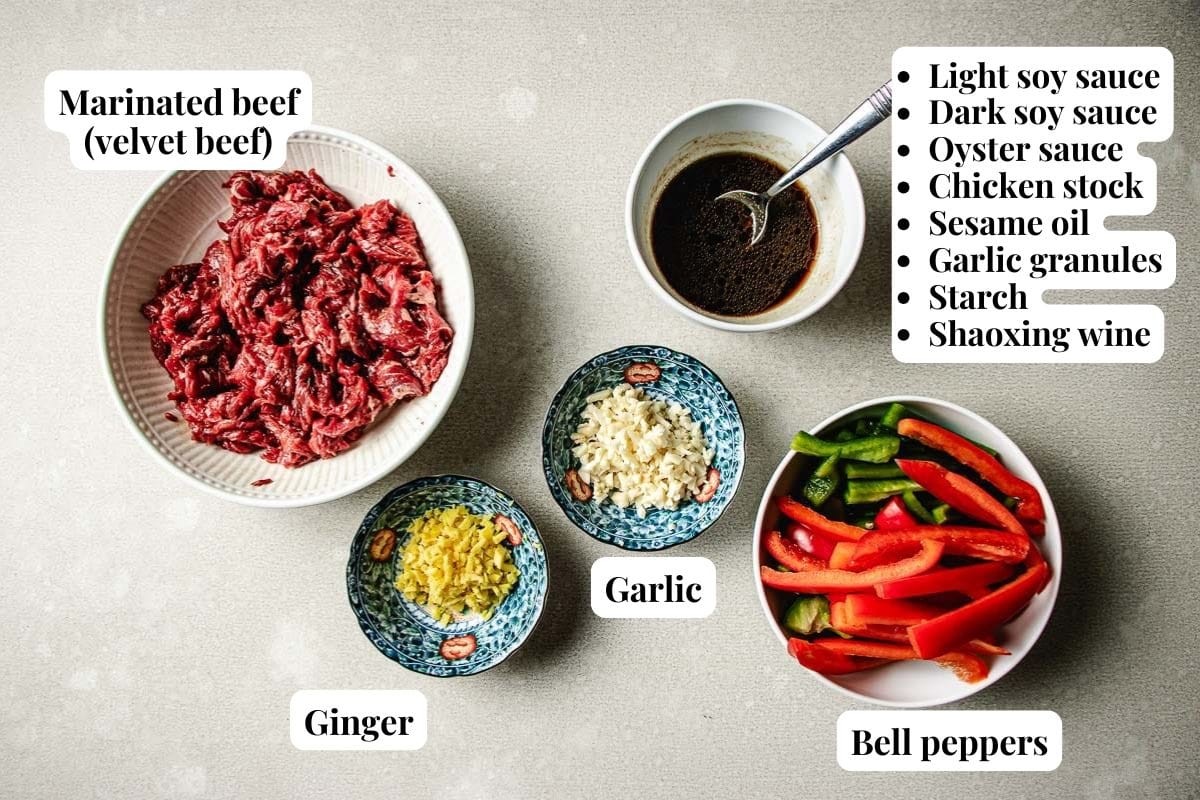 Five bowls display the essentials for an air fryer beef stir fry—marinated beef, minced garlic, minced ginger, sliced red and green bell peppers, and a dark marinade sauce—all with labeled ingredients arranged on a light surface.