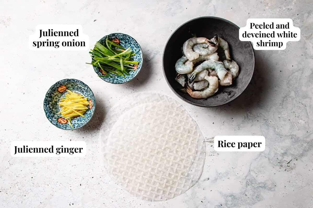 Overhead view of ingredients for rice paper shrimp chips: a bowl of peeled and deveined shrimp, julienned spring onion, julienned ginger, and a sheet of rice paper on a light surface, each labeled.