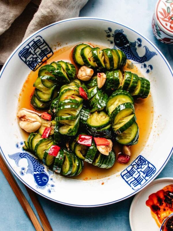 A plate of marinated cucumbers sliced and arranged in a spiral, garnished with red chili, garlic, and herbs, is served in a spicy sauce on a white dish with blue fish designs. Chopsticks and a sauce dish are placed beside the plate.