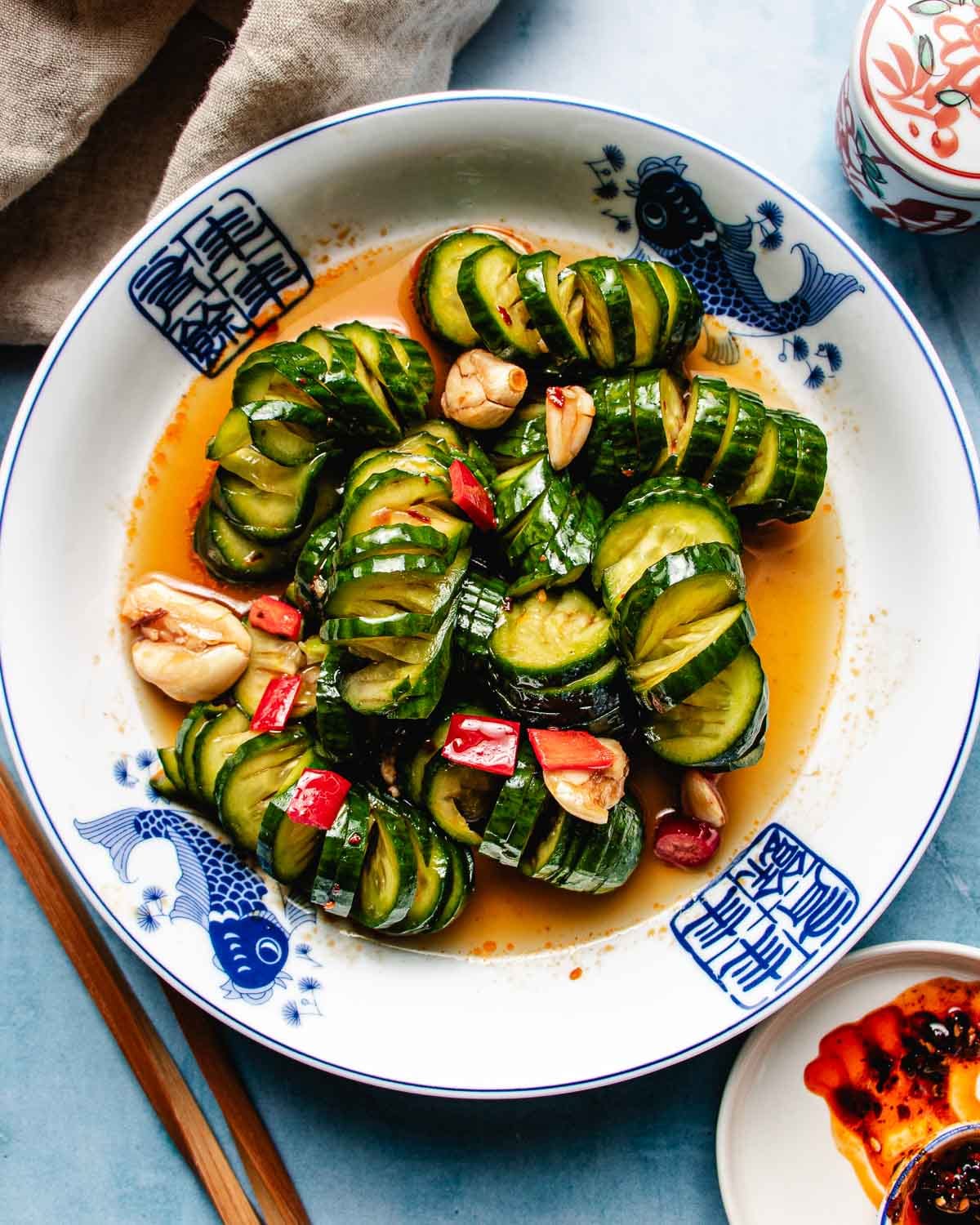 A plate of marinated cucumbers sliced and arranged in a spiral, garnished with red chili, garlic, and herbs, is served in a spicy sauce on a white dish with blue fish designs. Chopsticks and a sauce dish are placed beside the plate.