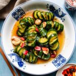 Marinated cucumbers are sliced and arranged in a bowl with garlic, red chili, and a savory sauce, then served on a blue and white plate with fish designs.