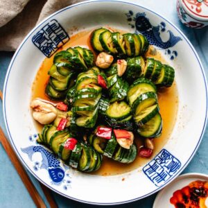 Marinated cucumbers are sliced and arranged in a bowl with garlic, red chili, and a savory sauce, then served on a blue and white plate with fish designs.