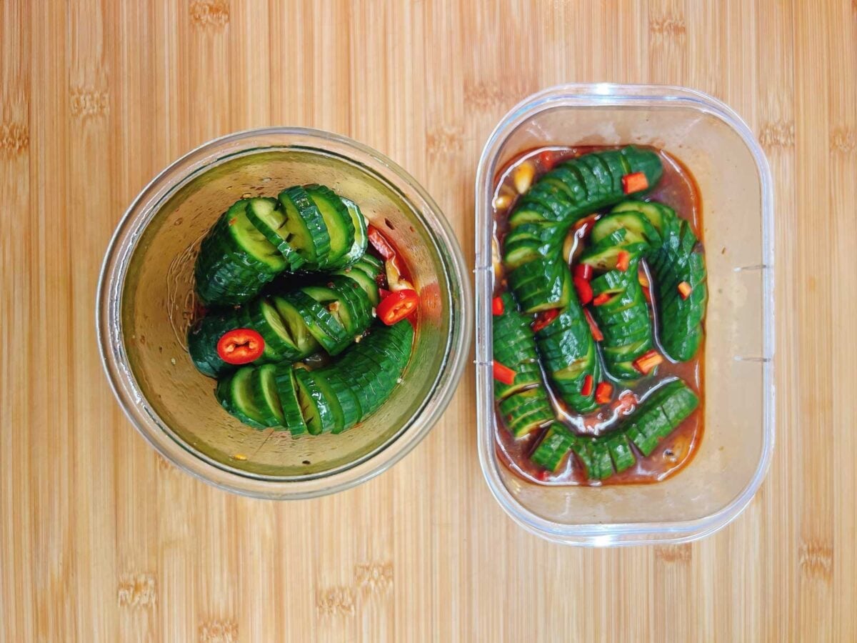 Two containers of marinated cucumbers in savory lightly sweet and tangy soy sauce marinade, garnished with red chili, are placed on a light wooden surface with cucumbers marinated in a glass jar and cucumbers marinated in a container.