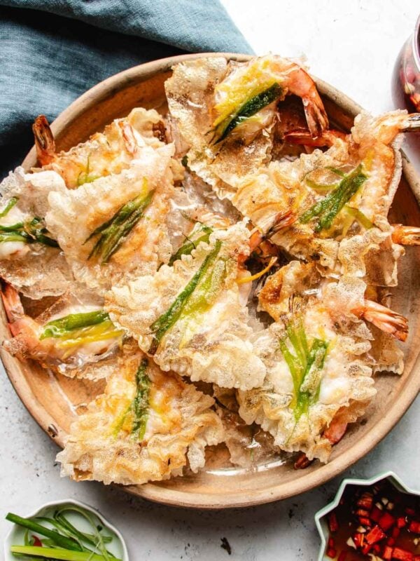 A wooden plate filled with crispy shrimp chips made with rice paper, each topped with a strip of green vegetable, sits on a light table next to small bowls of sliced green onions, chopped red chilies, and crunchy rice paper shrimp chips.