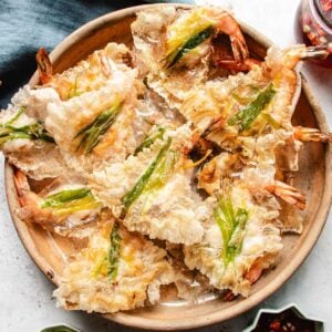 A wooden plate filled with crispy rice paper shrimp chips, topped with green chili slices. The golden-brown fritters are arranged in a slightly overlapping pattern and paired perfectly with crunchy shrimp chips.