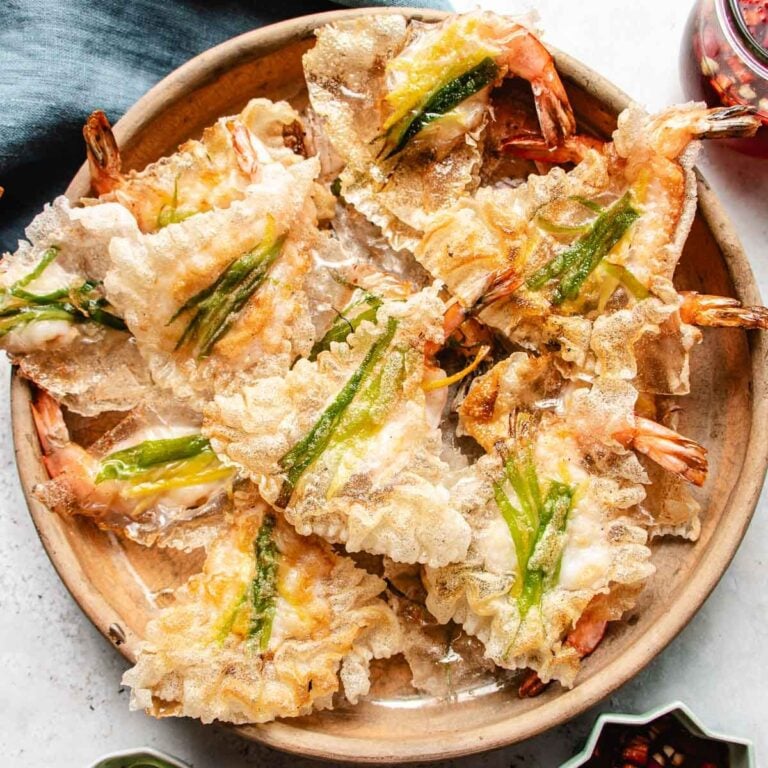 A wooden plate filled with crispy rice paper shrimp chips, topped with green chili slices. The golden-brown fritters are arranged in a slightly overlapping pattern and paired perfectly with crunchy shrimp chips.