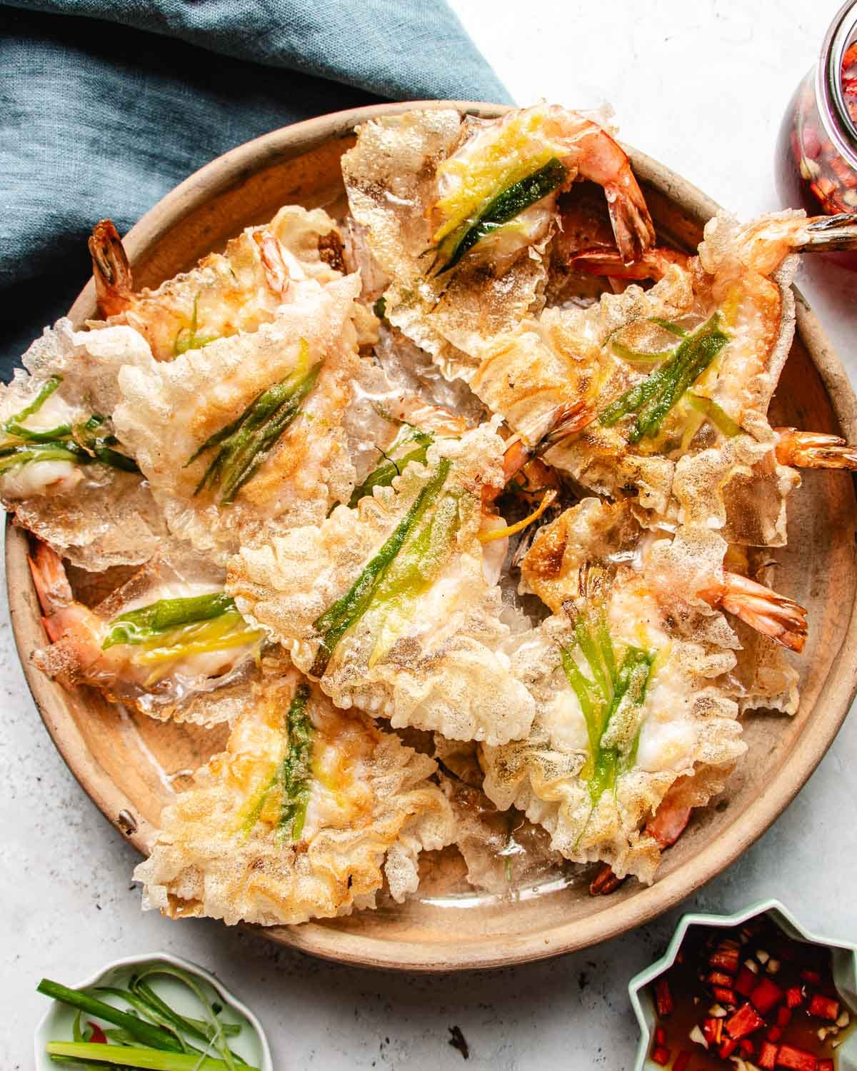A wooden plate filled with crispy shrimp chips made with rice paper, each topped with a strip of green vegetable, sits on a light table next to small bowls of sliced green onions, chopped red chilies, and crunchy rice paper shrimp chips.