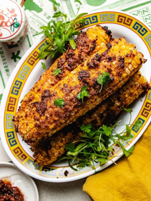 Four corn cobs coated with a savory reddish shacha sauce, reminiscent of Taiwanese street corn, are garnished with fresh cilantro and greens, served on a decorative plate with blue and yellow patterns. A yellow napkin and small bowl are nearby.