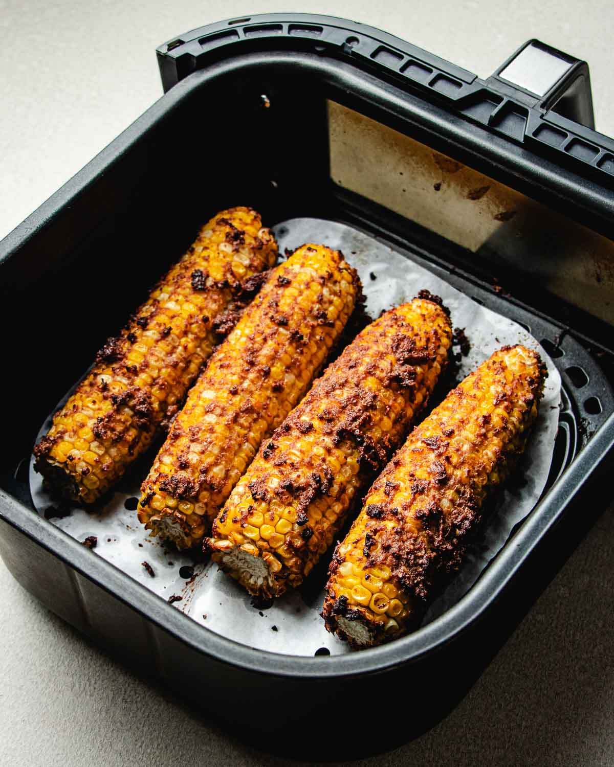 Four seasoned and roasted corn on the cobs, inspired by Taiwanese street corn, sit in an air fryer basket lined with parchment paper, showing a crispy, golden-brown coating.