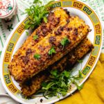Grilled Taiwanese street corn on the cob topped with a spicy seasoning and garnished with fresh cilantro, served on a decorative oval plate with a yellow patterned border.