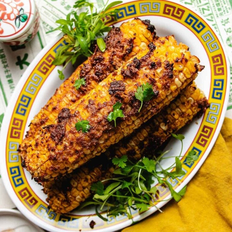 Grilled Taiwanese street corn on the cob topped with a spicy seasoning and garnished with fresh cilantro, served on a decorative oval plate with a yellow patterned border.