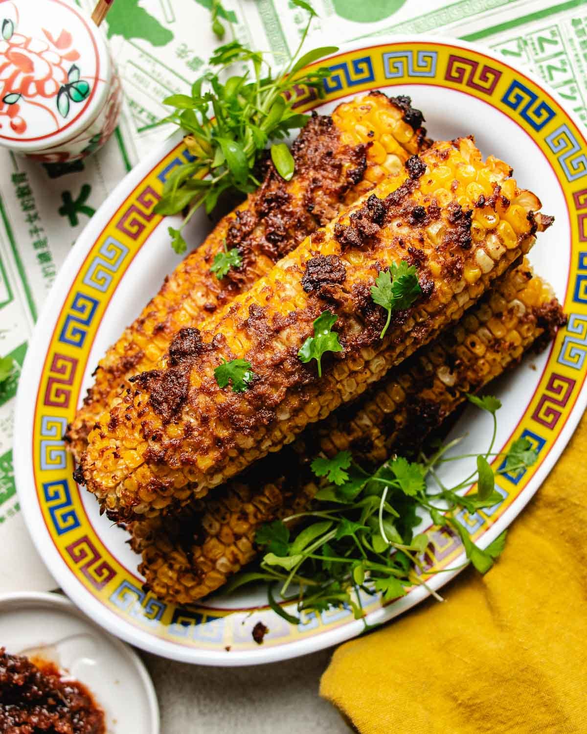 Four corn cobs coated with a savory reddish shacha sauce, reminiscent of Taiwanese street corn, are garnished with fresh cilantro and greens, served on a decorative plate with blue and yellow patterns. A yellow napkin and small bowl are nearby.