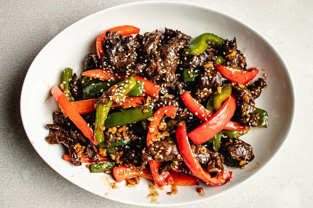A white plate filled with air fryer beef stir fry, featuring tender strips of beef, red and green bell peppers, carrots, and sesame seeds in a savory sauce.