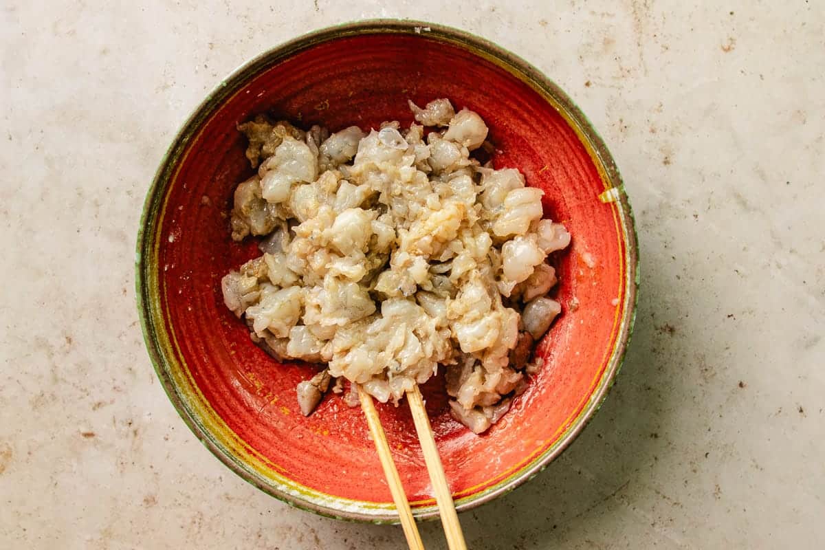 A red bowl filled with finely chopped raw shrimp, being mixed with wooden chopsticks, sits on a light-colored countertop—perfectly prepared as a filling for delicious shrimp lettuce wraps.