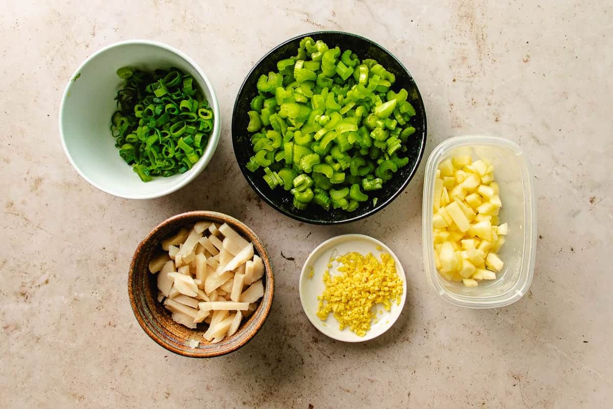 Five bowls with chopped green onions, celery, diced apples, crunchy wonton strips, and minced ginger on a light countertop—perfect fresh ingredients for making flavorful shrimp lettuce wraps.