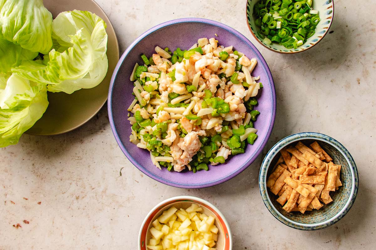 A bowl of diced and cooked shrimp with vegetables is surrounded by lettuce wraps, chopped green onions, fried wonton strips, and diced apples on a light surface—perfect for assembling flavorful shrimp lettuce wraps.