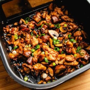 Sliced air fryer chicken stir fry and mushrooms in a dark sauce, topped with sliced green onions, in a black rectangular dish on a wooden surface.