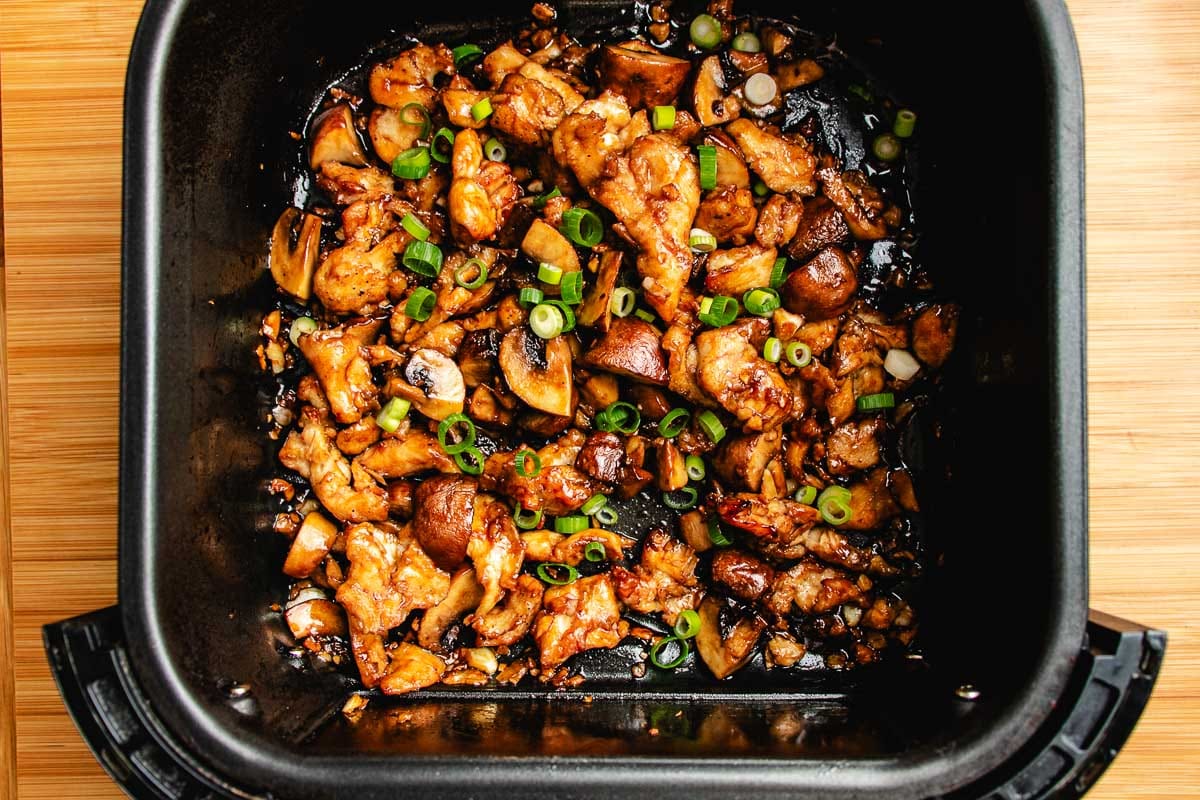 Cooked air fryer chicken stir fry pieces with mushrooms and chopped green onions in a black air fryer basket on a wooden surface.