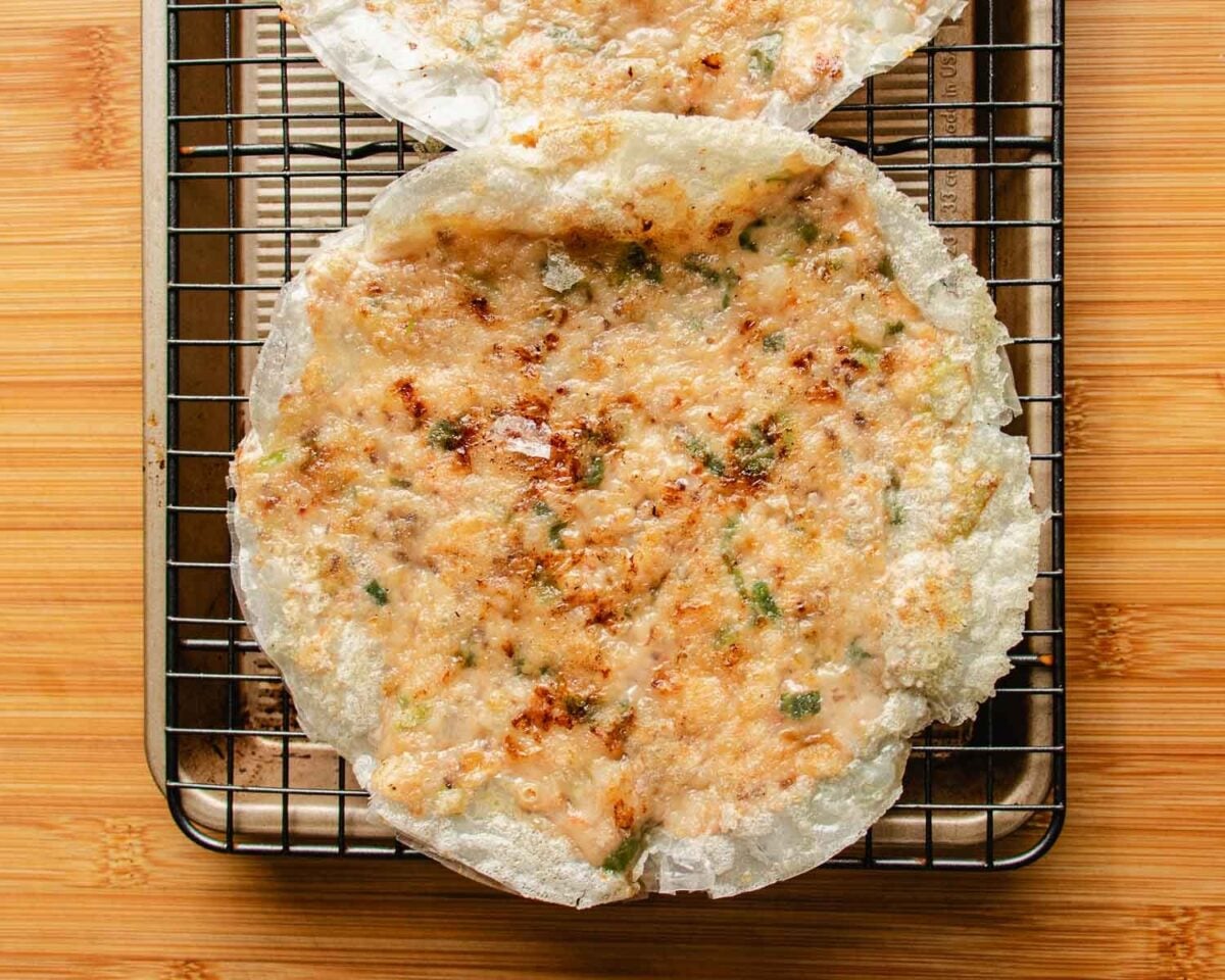 A crispy Vietnamese rice paper shrimp pancake snack topped with scallions and browned, melted cheese sits on a cooling rack over a baking sheet, on a wooden surface.