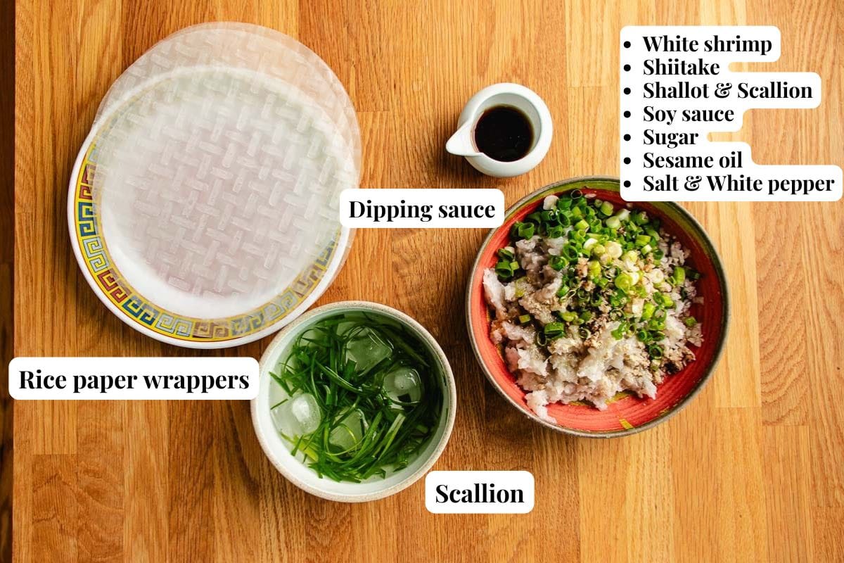 Rice paper wrappers, a bowl of scallions in water, a bowl filled with shrimp and mushrooms topped with scallion—perfect for making rice paper shrimp pancakes—a small dish of dipping sauce, and labeled ingredient notes are artfully arranged on a wooden table.