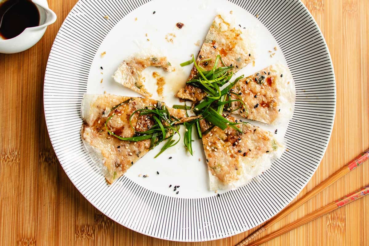 Six triangular slices of savory rice paper shrimp pancakes topped with green onions, sesame seeds, and sauce are arranged on a white plate with black lines, with chopsticks and a small cup of soy sauce on the side.