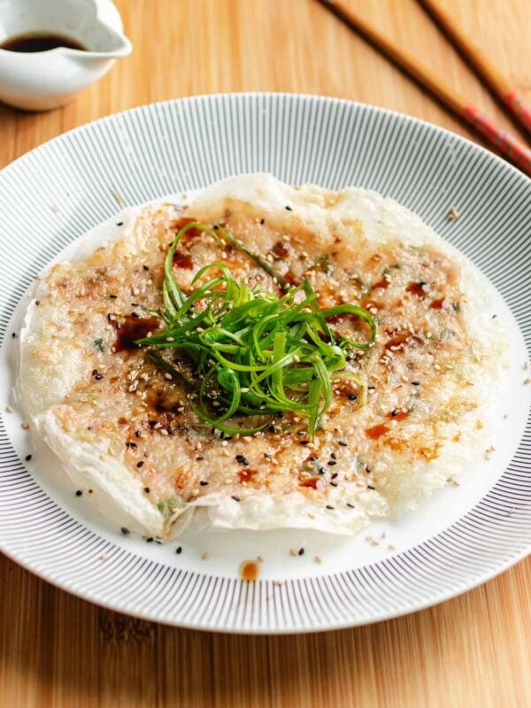 A plate of crispy rice paper shrimp pancakes topped with green onions, sesame seeds, and sauce, served on a striped plate with chopsticks and a small bowl of sauce in the background.