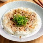 A crispy rice paper pancake with a golden crust, reminiscent of traditional rice paper shrimp pancakes, topped with sliced green onions, sesame seeds, and a drizzle of sauce, served on a white plate with a patterned rim.