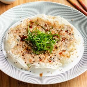 A crispy rice paper pancake with a golden crust, reminiscent of traditional rice paper shrimp pancakes, topped with sliced green onions, sesame seeds, and a drizzle of sauce, served on a white plate with a patterned rim.