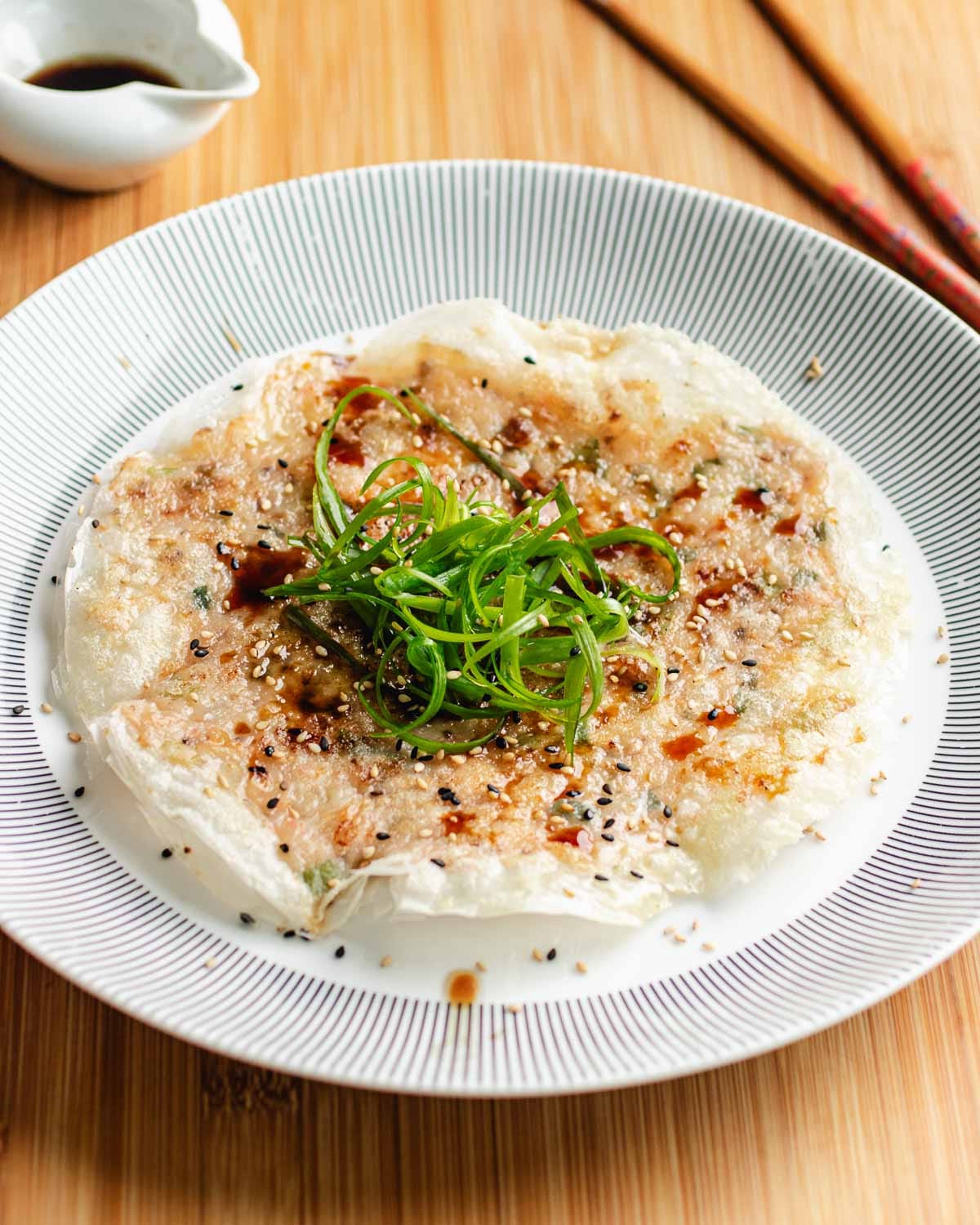 A plate of crispy rice paper shrimp pancakes topped with green onions, sesame seeds, and sauce, served on a striped plate with chopsticks and a small bowl of sauce in the background.