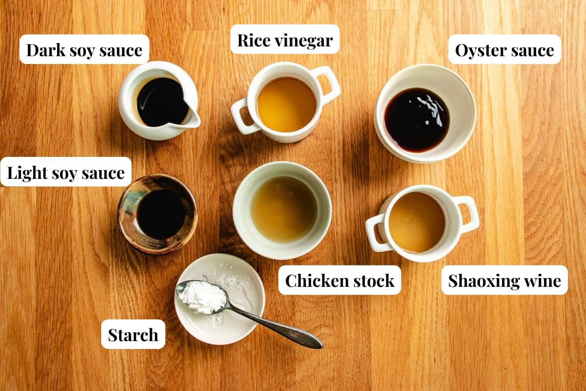 Seven small bowls labeled with ingredients—dark soy sauce, light soy sauce, rice vinegar, chicken stock, oyster sauce, starch (with a spoon), and Shaoxing wine—are all arranged on a wooden surface, ready for making a flavorful Air fryer chicken stir fry.