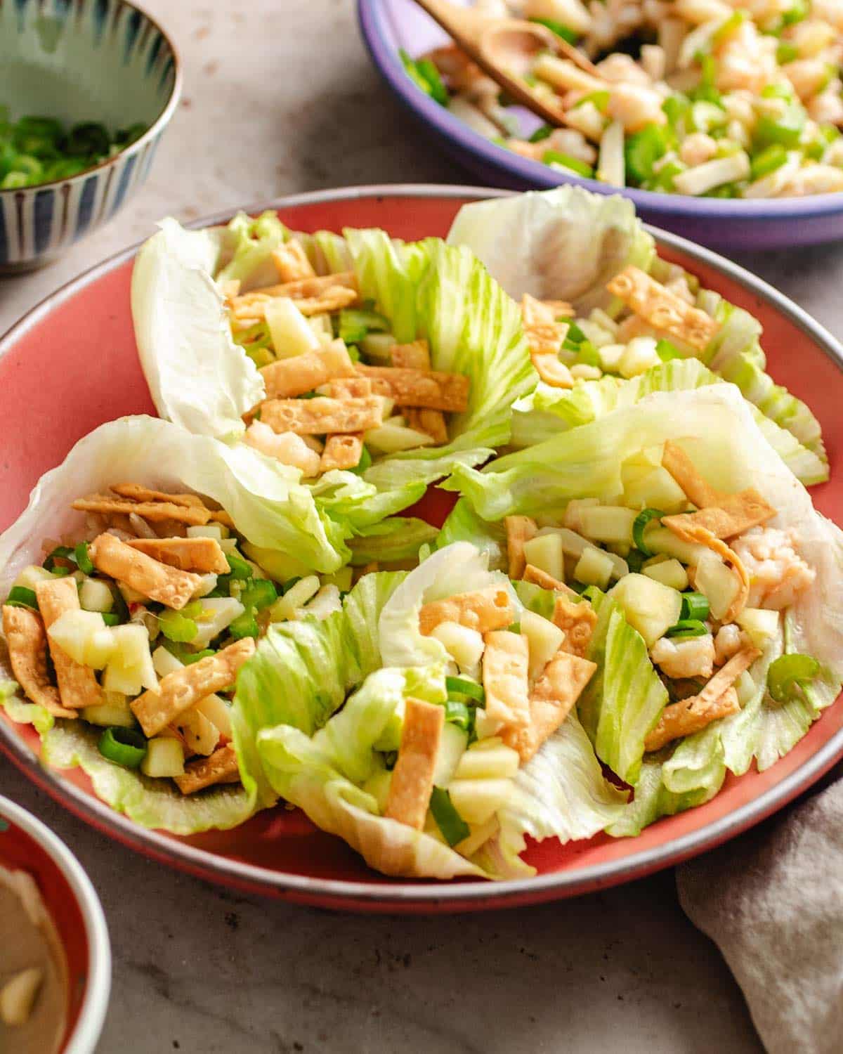 Six lettuce leaves filled with chopped and cooked shrimp, diced vegetables, and crispy wonton strips are arranged on a red plate, offering a Taiwanese-style shrimp lettuce wraps. Bowls with more ingredients are in the background.