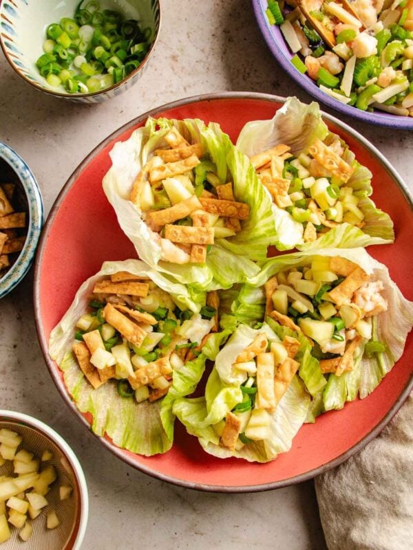 Four lettuce cups—similar to classic shrimp lettuce wraps—filled with diced apples, wonton strips, and green onions on a red plate, surrounded by bowls of extra ingredients.