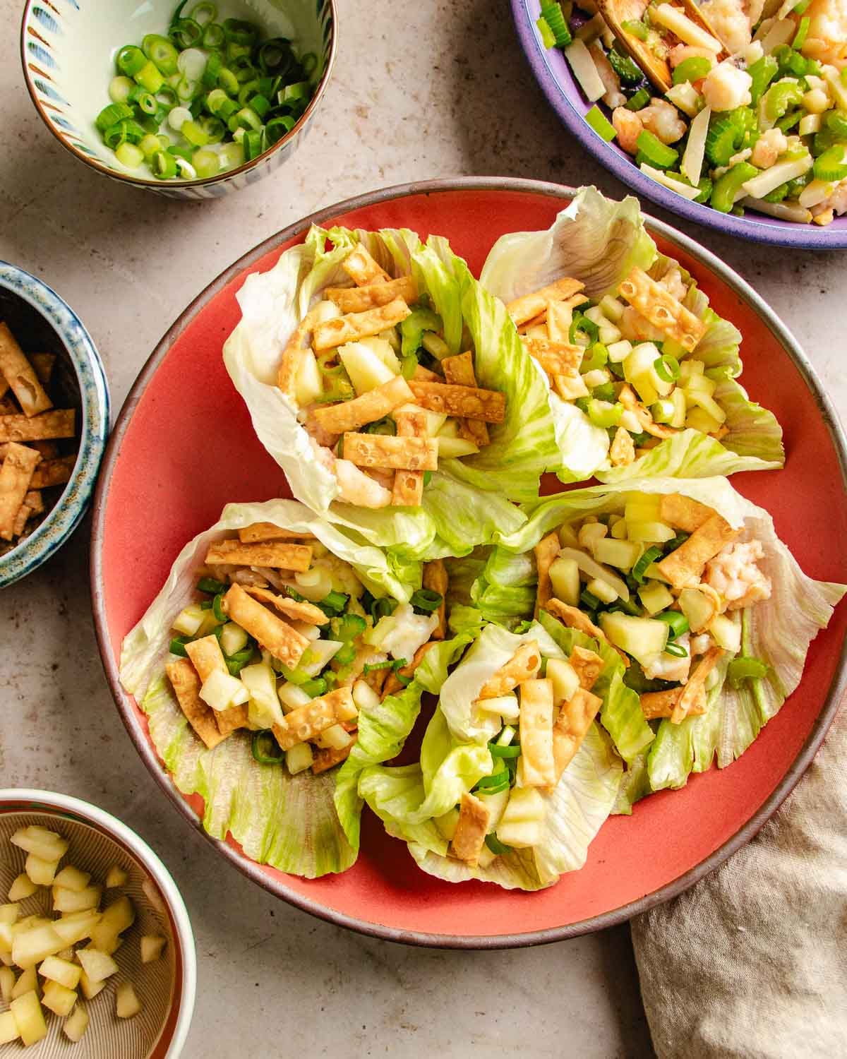 Four lettuce cups—similar to classic shrimp lettuce wraps—filled with diced apples, wonton strips, and green onions on a red plate, surrounded by bowls of extra ingredients.