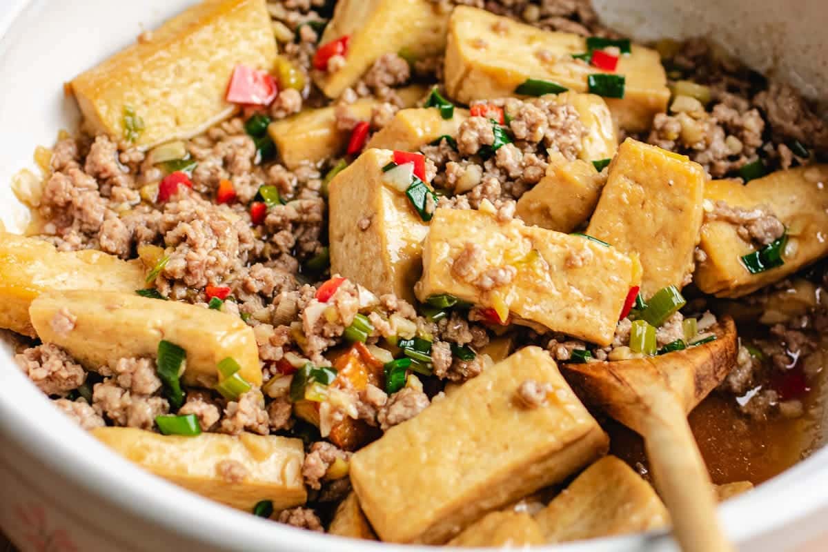 Pan fried tofu with minced pork, chopped green onions, and red peppers in a light sauce, served in a white bowl with a wooden spoon.