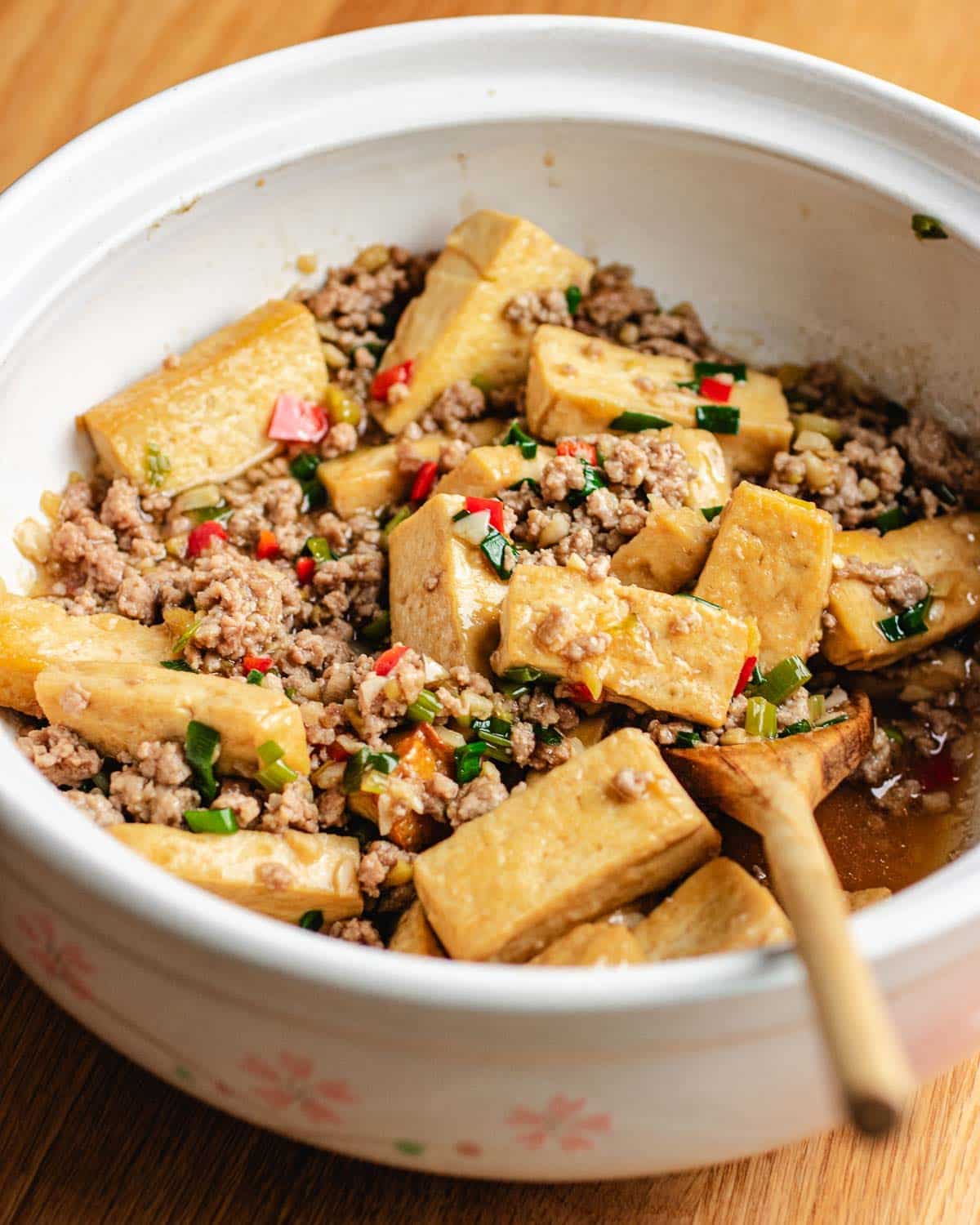 A ceramic bowl filled with tofu cubes, minced pork, chopped green onions, and red chili, with a wooden spoon for serving—perfect for enjoying classic tofu with minced pork.