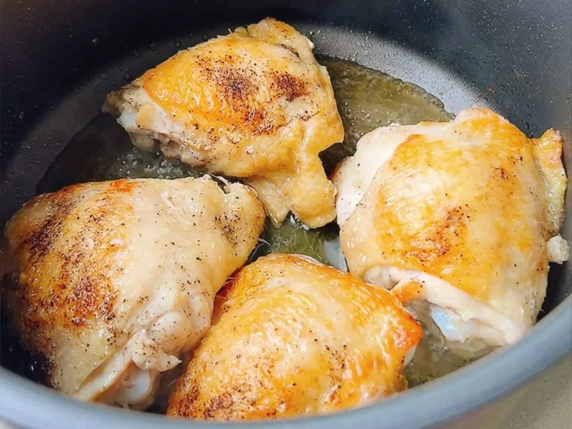 Four golden-brown chicken thighs are being seared in a pan, with visible oil and seasoning on the crispy skin—the perfect base for a savory Shiitake chicken soup.