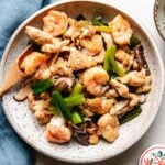 A speckled plate filled with Chicken and Shrimp Stir Fry—tender stir-fried shrimp, sliced chicken, mushrooms, and green onions—rests beside a wooden spoon and a blue napkin.