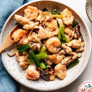 A speckled plate filled with Chicken and Shrimp Stir Fry—tender stir-fried shrimp, sliced chicken, mushrooms, and green onions—rests beside a wooden spoon and a blue napkin.