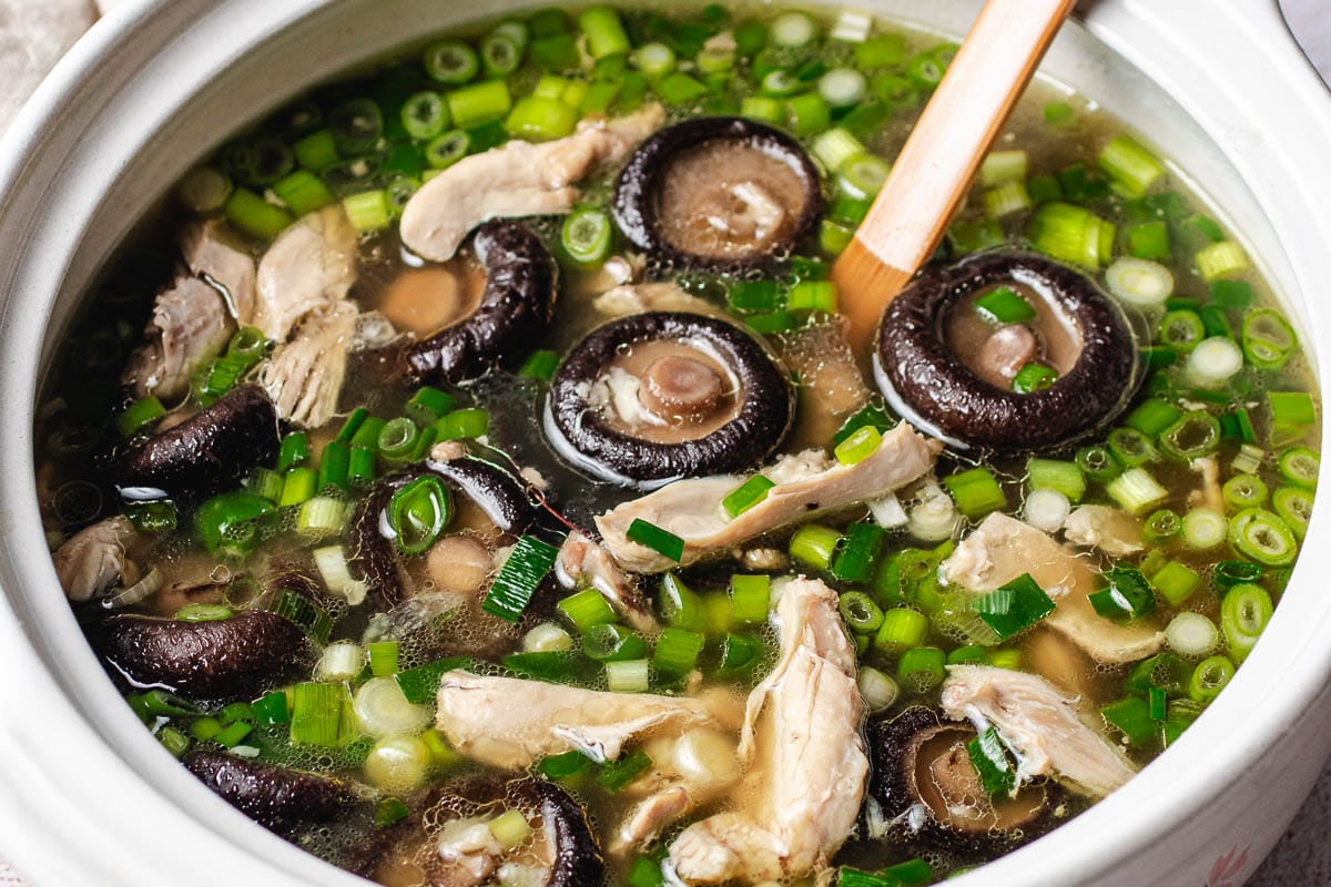 A pot of Chinese chicken mushroom soup with chicken pieces, sliced shiitake mushrooms, and chopped green onions, with a wooden spoon resting inside the pot.