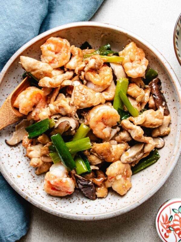 A ceramic plate holds a delicious Chicken and Shrimp Stir Fry, featuring tender shrimp, chicken, mushrooms, and green onions. A wooden spoon rests in the food, and a blue napkin is beside the plate.