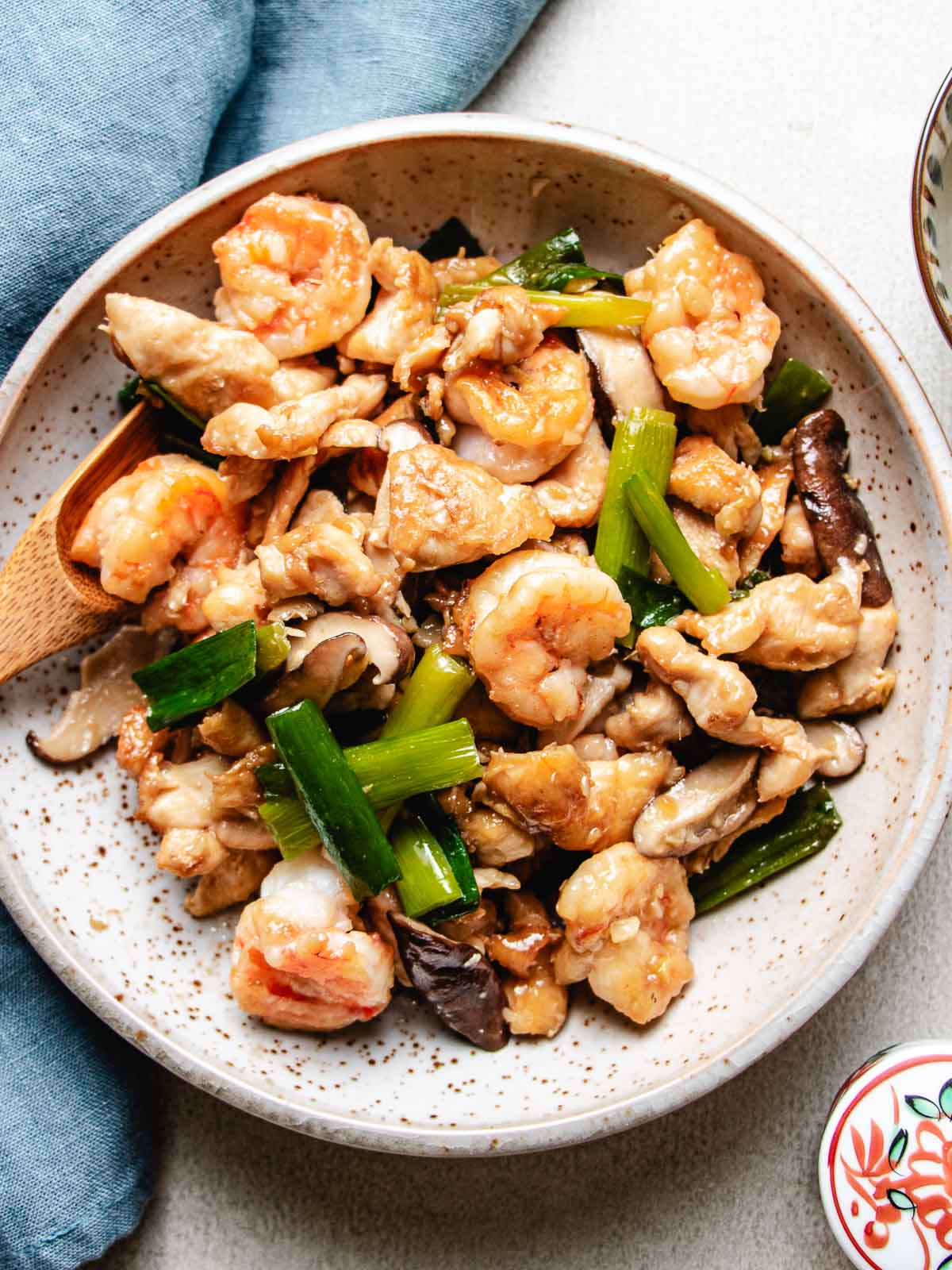 A ceramic plate holds a delicious Chicken and Shrimp Stir Fry, featuring tender shrimp, chicken, mushrooms, and green onions. A wooden spoon rests in the food, and a blue napkin is beside the plate.