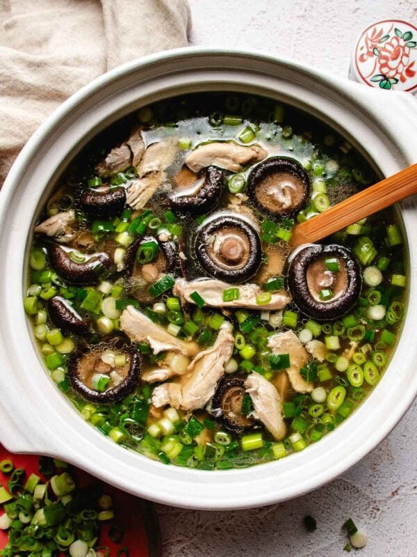 A bowl of Chicken shiitake mushroom soup with sliced mushrooms, shredded chicken, and chopped green onions, a wooden spoon resting inside. A plate of green onions and a beige napkin are nearby.