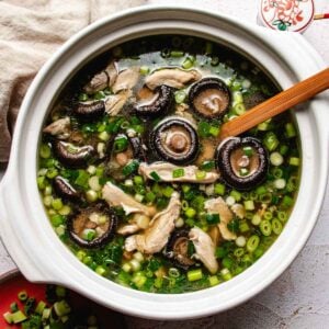 A ceramic pot filled with Chinese chicken mushroom soup containing sliced shiitake, shredded chicken, and chopped green onions, with a wooden spoon resting inside.