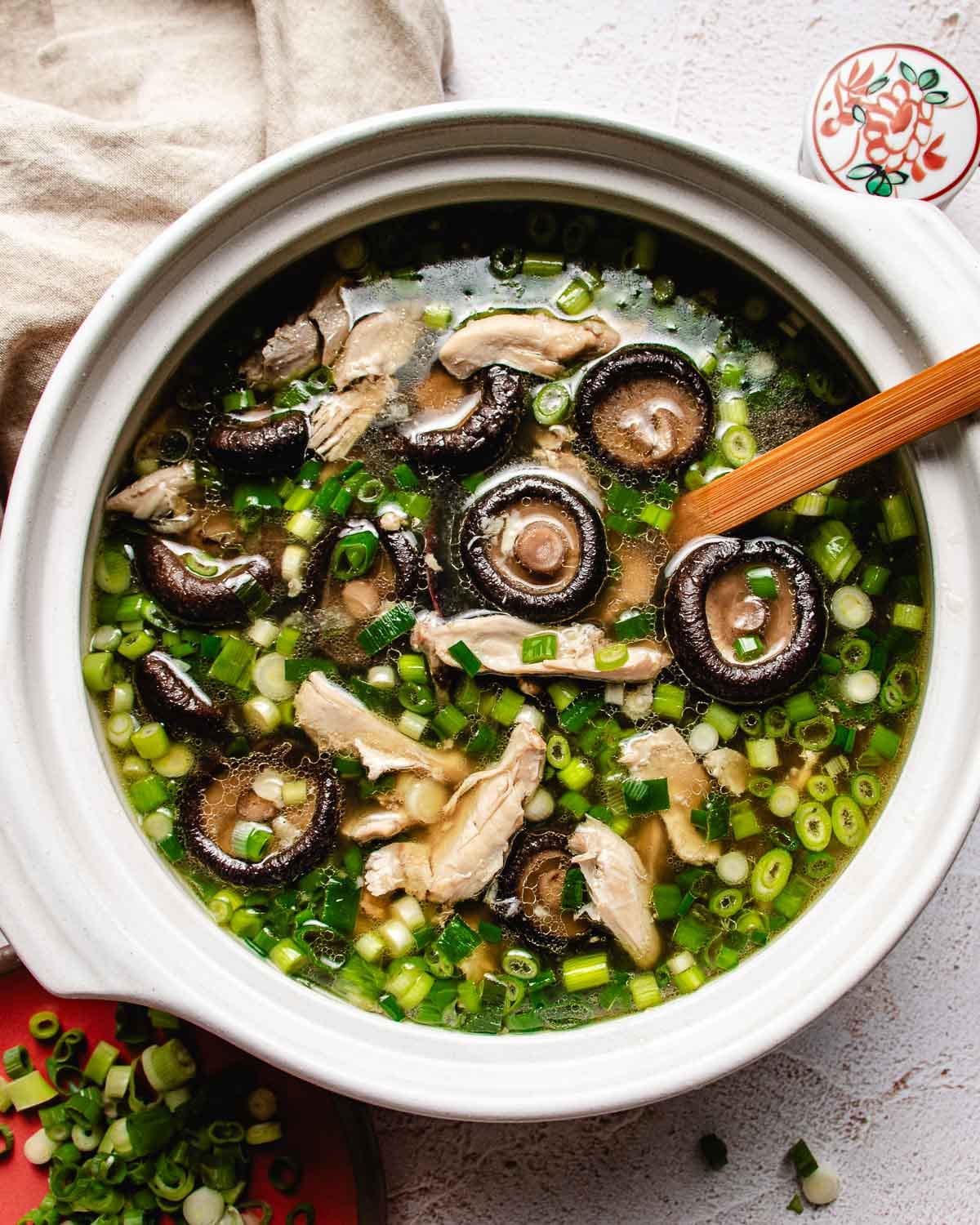A bowl of Chicken shiitake mushroom soup with sliced mushrooms, shredded chicken, and chopped green onions, a wooden spoon resting inside. A plate of green onions and a beige napkin are nearby.