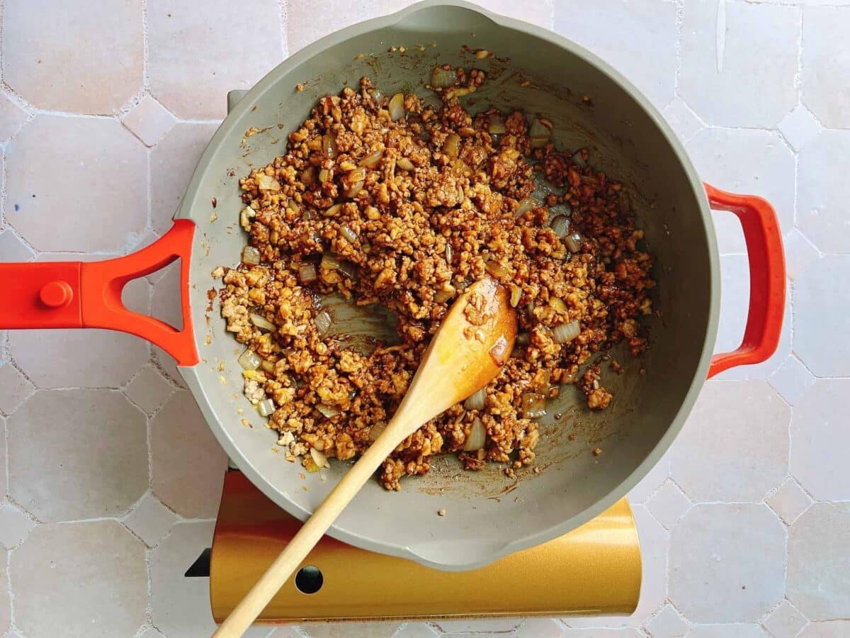 A pan with a red handle holds cooked ground meat and diced vegetables, being stirred with a wooden spoon on a light tiled surface—a delicious filling that would be perfect for a wonton wrapper noodle bowl.