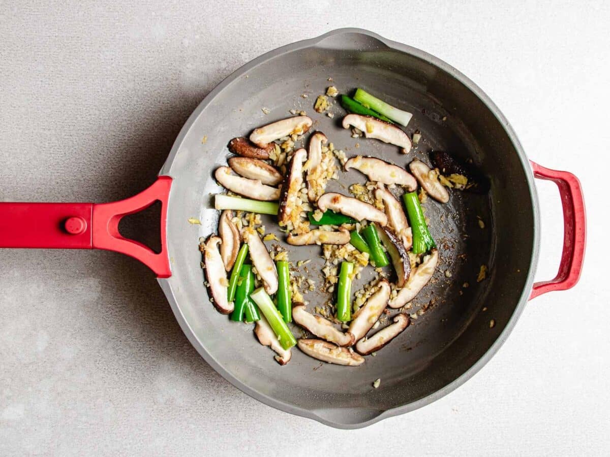 A gray skillet with red handles contains sautéed sliced shiitake and chopped green onions on a light surface, perfect as a flavorful base for a Chicken and Shrimp Stir Fry.