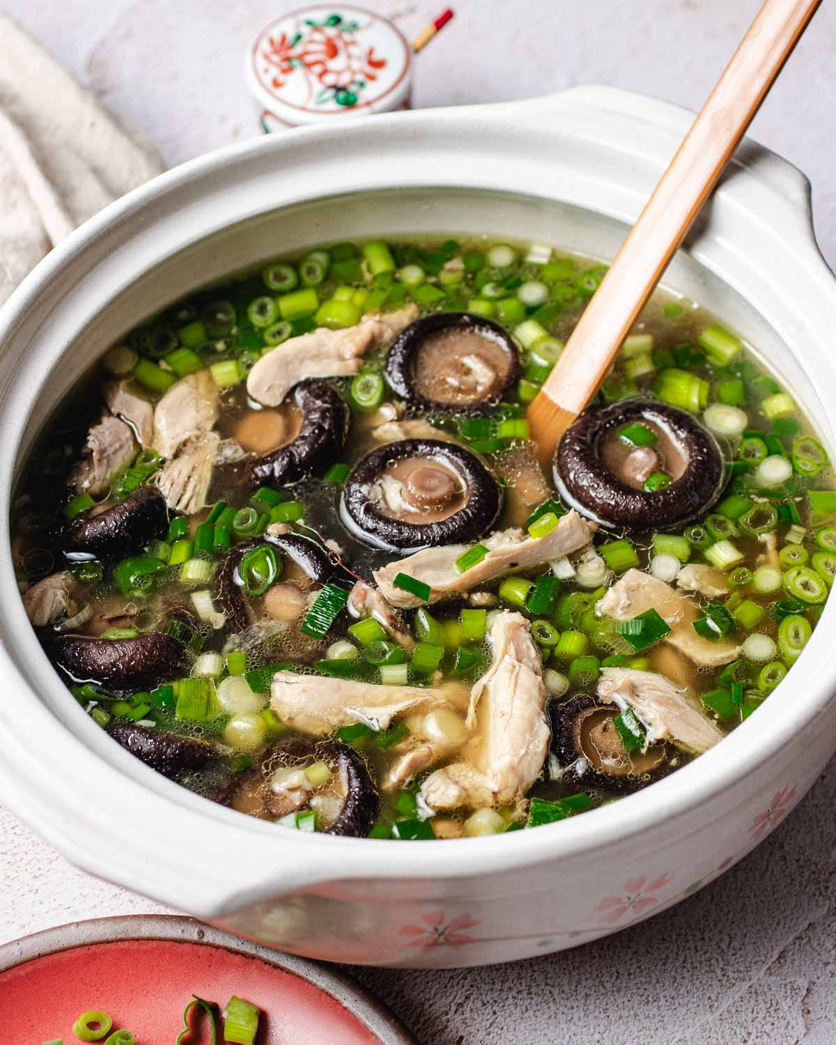 A ceramic pot filled with Chinese chicken mushroom soup—香菇雞湯—featuring tender chicken and shiitake, garnished with green onions. A wooden ladle rests in the soup, a small dish sits nearby.