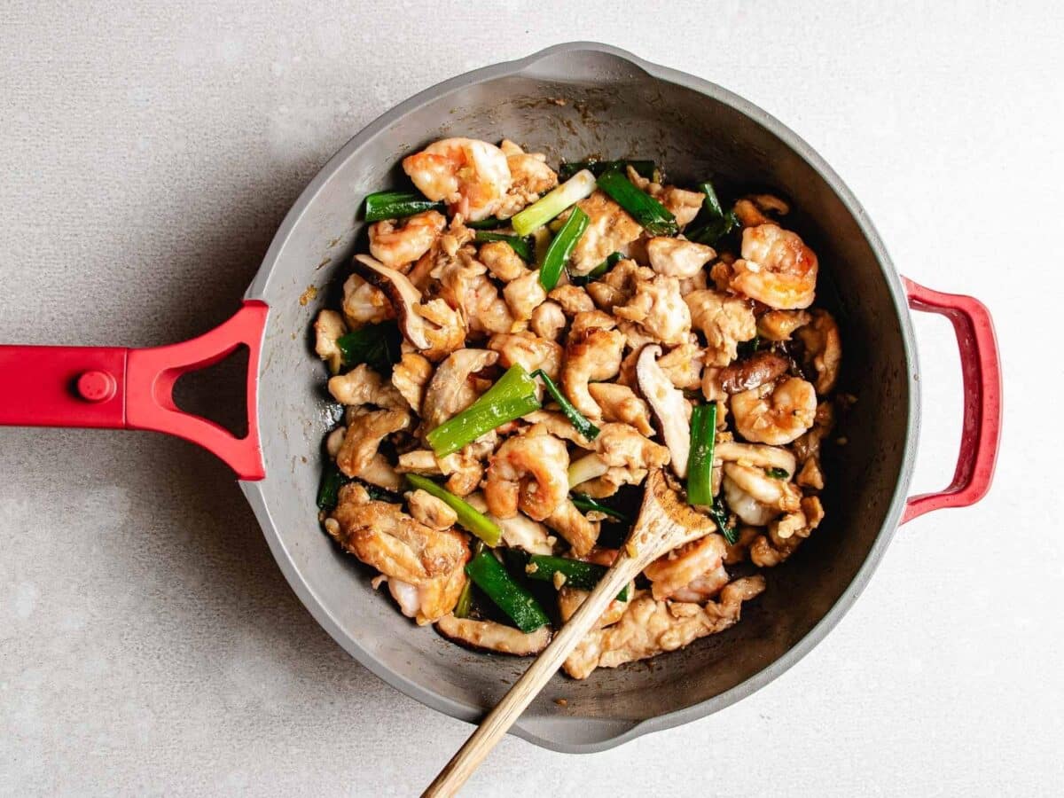 A pan with Chicken and Shrimp Stir Fry, featuring tender chicken, succulent shrimp, mushrooms, and green onions being mixed with a wooden spoon.