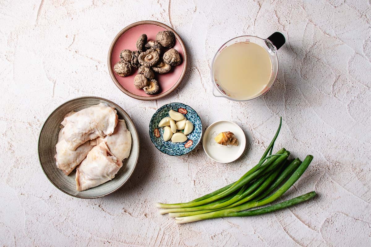Raw chicken, dry shiitake mushrooms, green onions, garlic and ginger, spices, and broth are arranged on a light textured surface—perfect ingredients for Chinese chicken mushroom soup.
